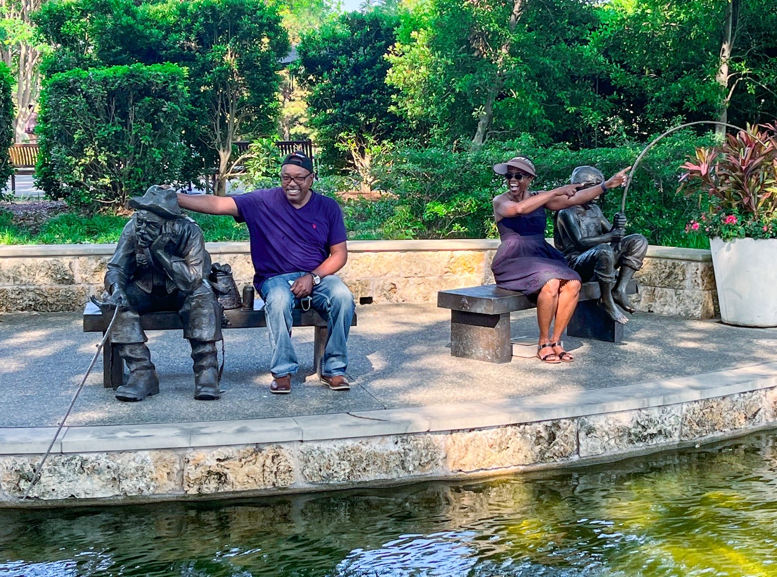 A man and a woman sit on separate benches beside a sculpture—she next to a young boy fishing, he beside an old man fishing—both laughing and pointing as they appreciate the scene, showing why Dallas is worth visiting