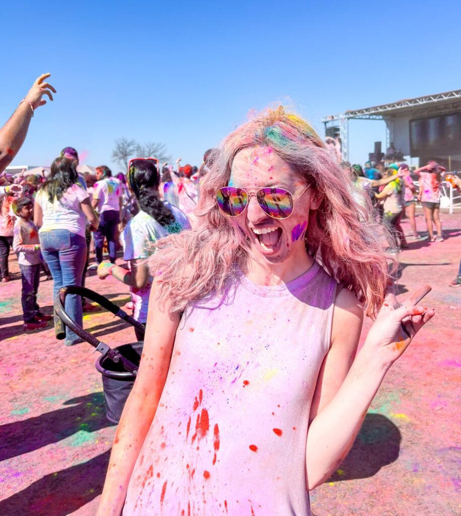 A young lady is standing, covered in pink chalk and smiling widely as the wind whips her long blond hair at the Holi Dallas Festival