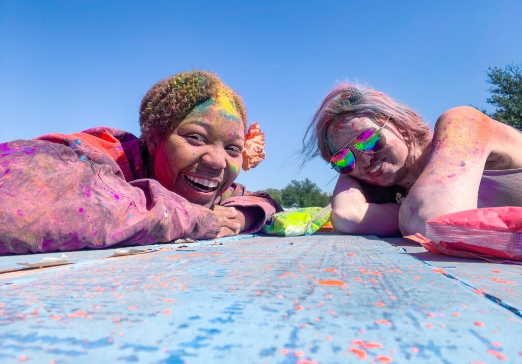 Two young ladies lying on the ground covered with blue, yellow, pink and green chalk, smiling contently at the Holi Dallas Festival