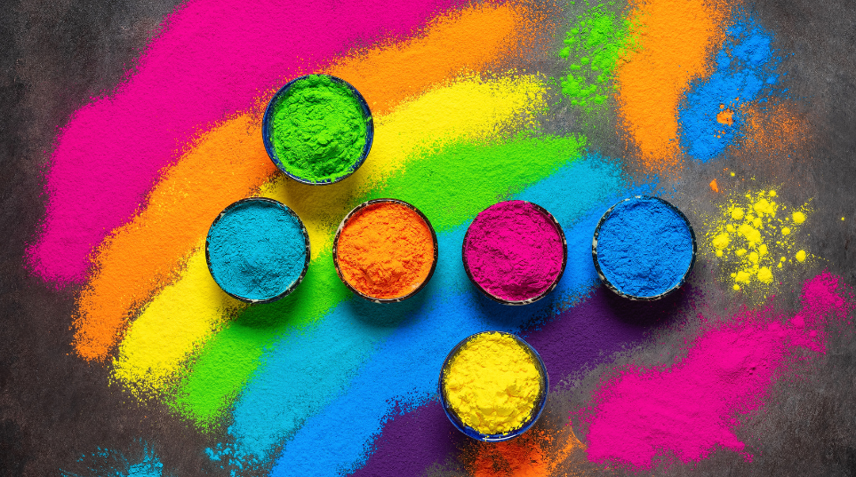 There are six pots of vibrant colors sitting on the pavement.  Aqua Blue, Electric Blue, Orange, Dark Pink, Yellow and Pea Green. These colors are common at the Holi Dallas Festival.