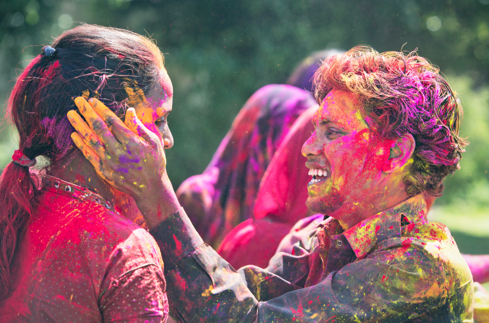 A young man frames the face of a young woman as they laugh and are covered with pink and yellow chalk at the Holi Dallas Festsival