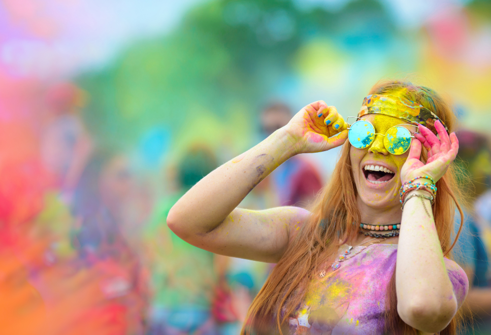 Young lady holds her hands up to steady her sunglasses as she laughs in delight at the Holi Dallas Festival