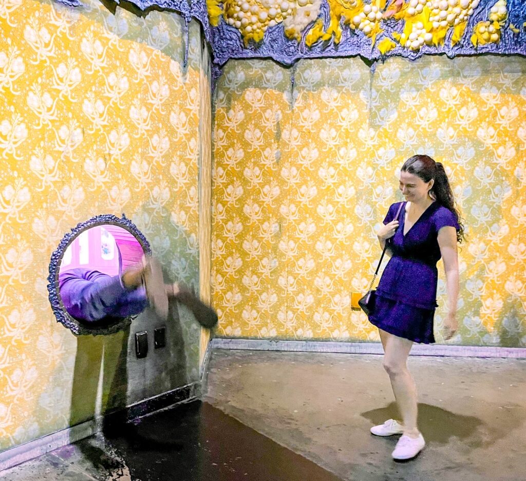 Young lady in a dress laughs as her friend's feet dangle from a wall opening at Meow Wolf during indoor activities in Dallas