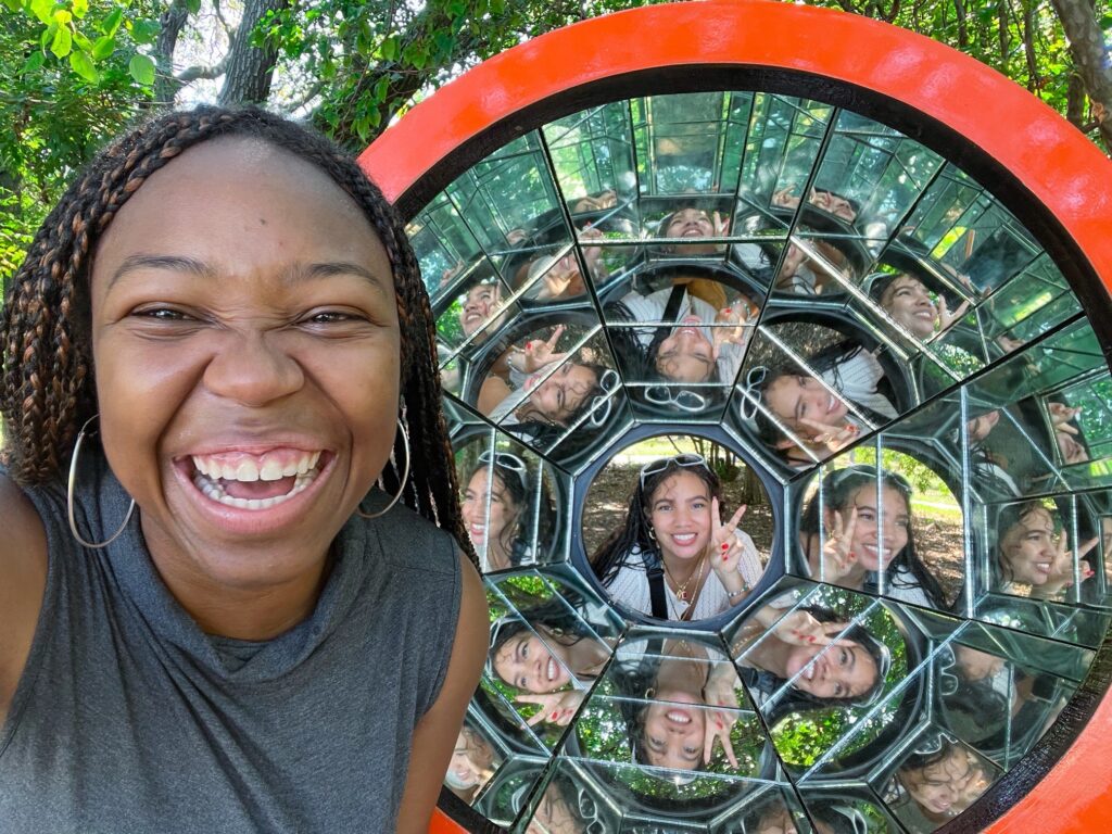 A young woman laughs as her friend’s reflection appears in a kaleidoscope-style mirrored public art installation, highlighting one of the unique-things-to-do-in-dfw.