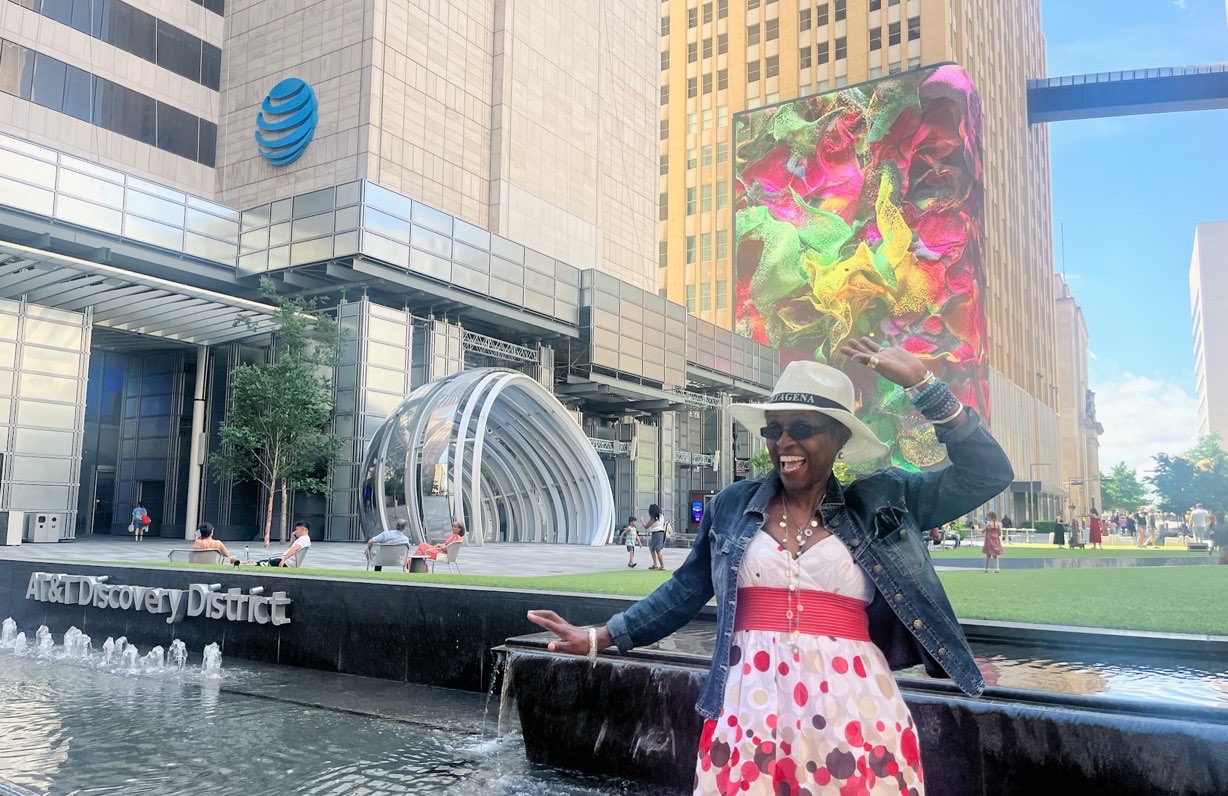 A lady smiles and poses in front of AT&T Discovery District. There is a beautiful multicolored abstract mural painted on a building in the background, part of free things to do in Dallas at night.