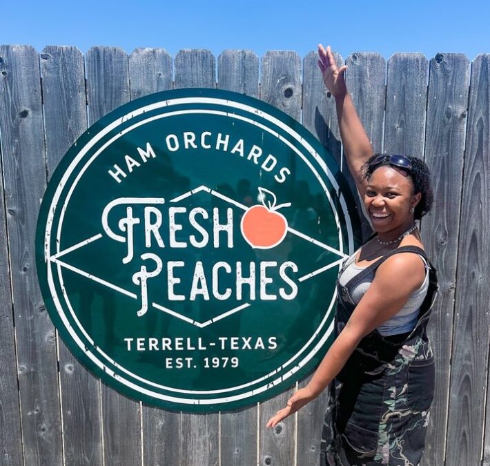 a young lady is standing next to a sign advertising fresh peaches at a Peach farm near Dallas. is dallas worth visiting with kids? Absolutely, yes.
