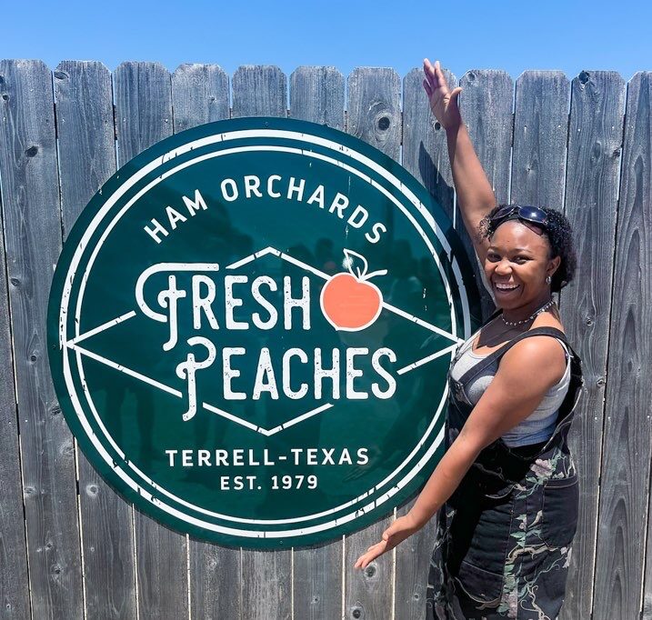 a young lady is standing next to a sign advertising fresh peaches at a Peach farm near Dallas. is dallas worth visiting with kids? Absolutely, yes.