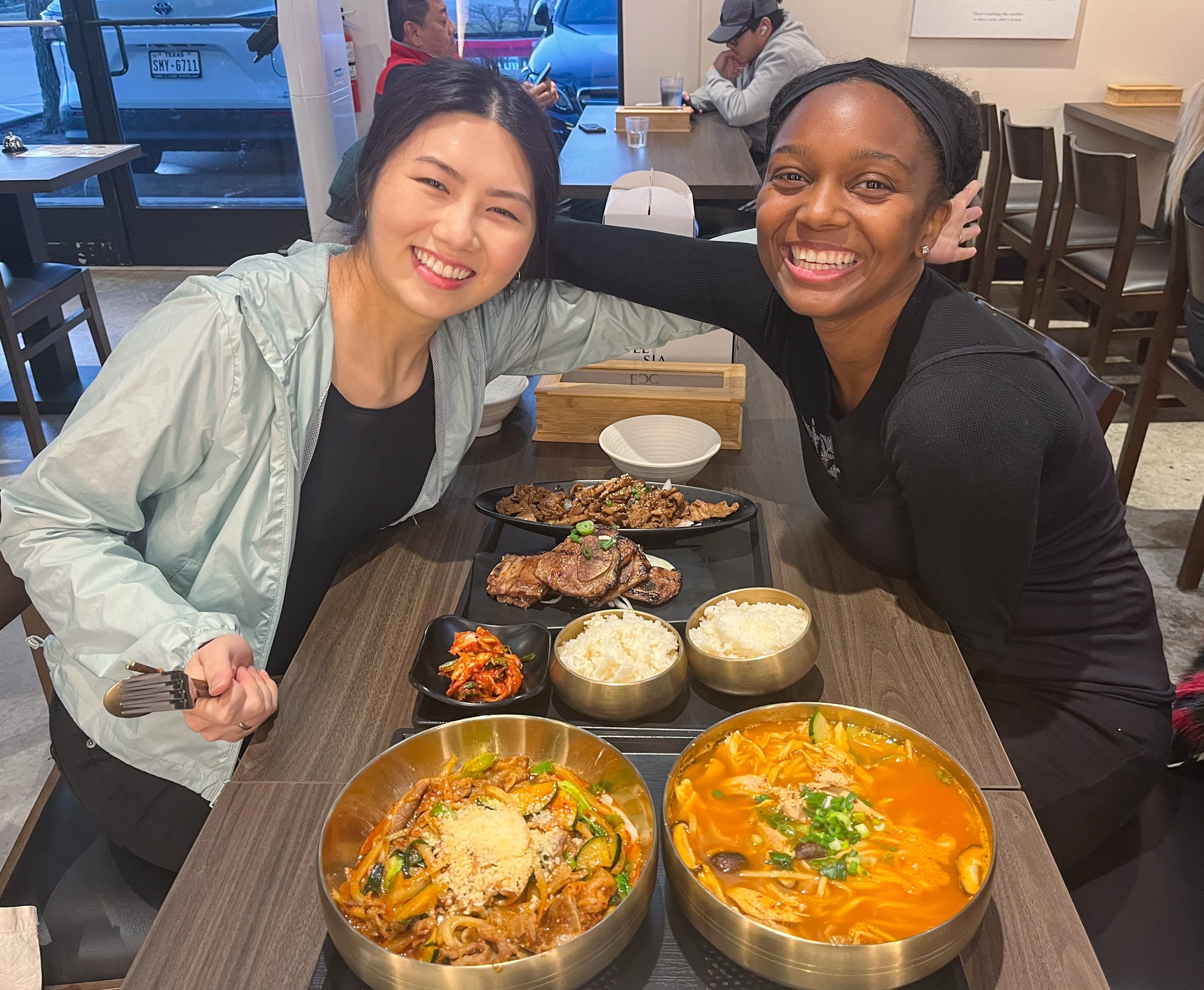 Friends pose before a Korean feast at Kooksoo Korean Restaurant in Plano, with noodles, rice, kimchi, soup, lamb chops, and chicken on the table, a unique-things-to-do-in-dfw experience.