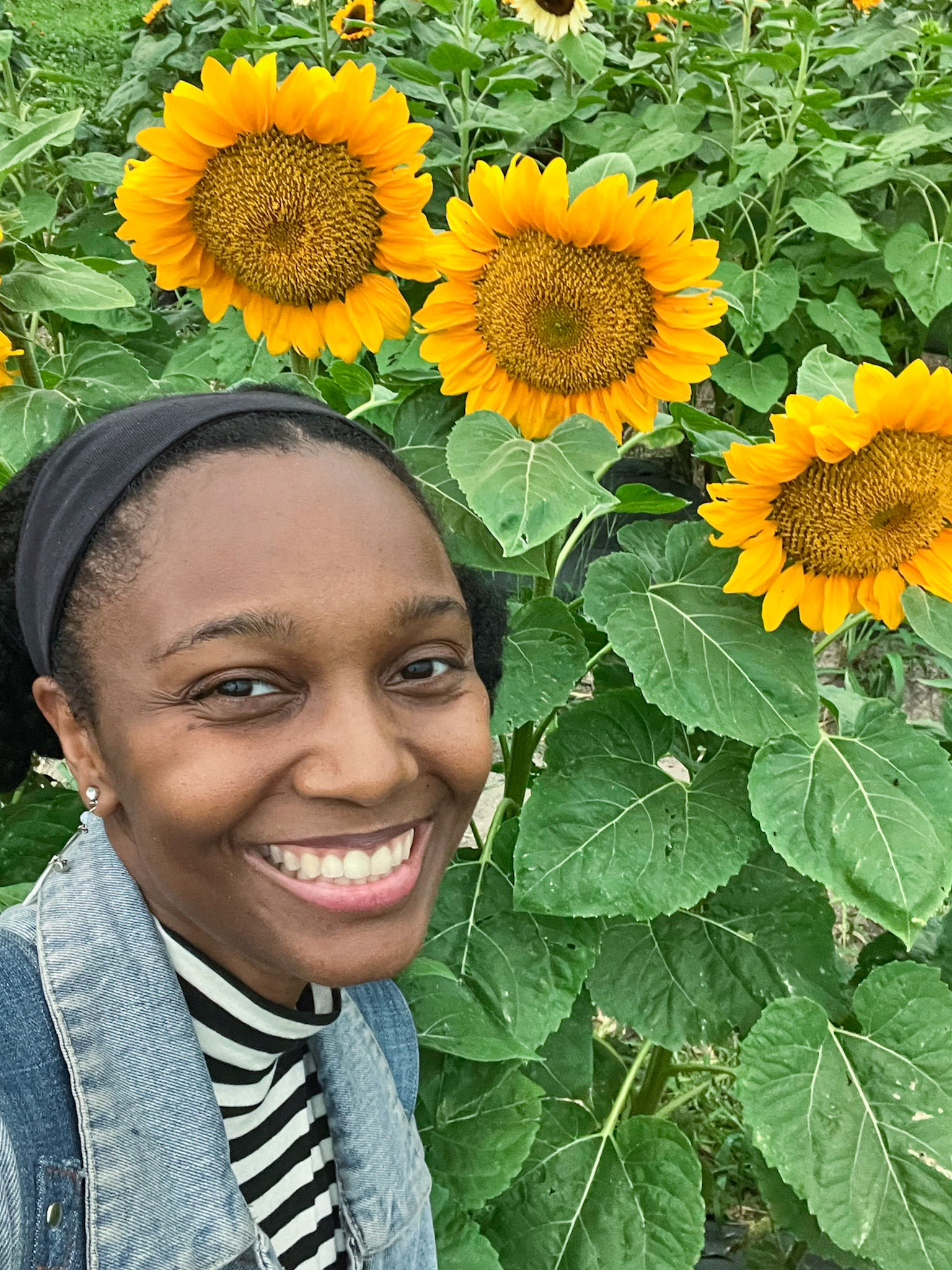 A woman smiling at the camera while squatting beside sunflowers.