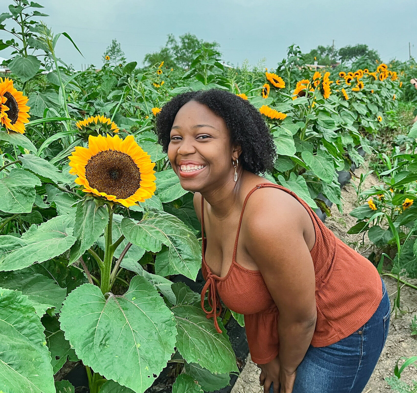 Young woman smiling beside a large sunflower in a sunflower field at one of the Dallas farms.