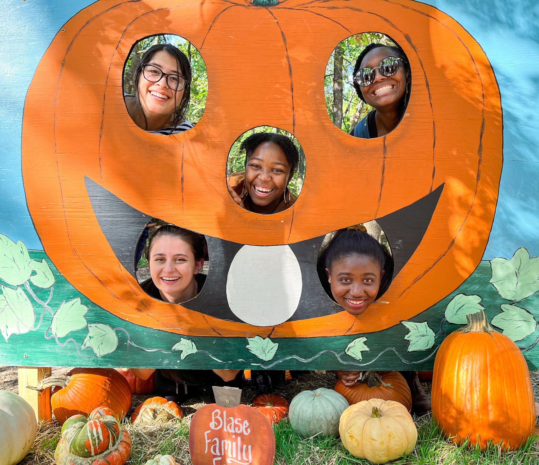 Five young ladies take photos with their faces in a large pumpkin cutout display at Blase Family Farm, one of the Dallas Farms near Dallas, Texas; the cutout has two eyes, a nose, and three teeth, with the middle front tooth slot empty.
