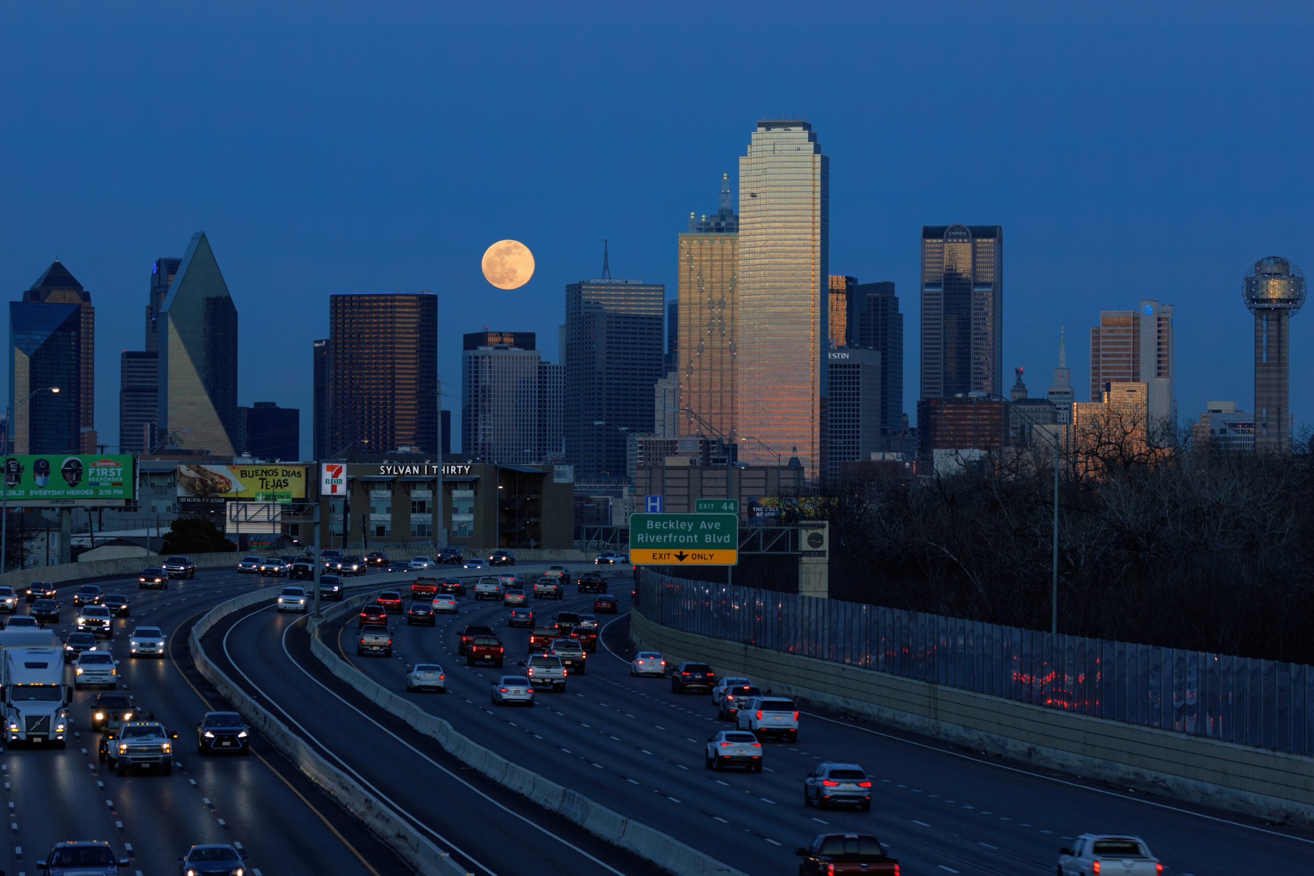 Cars stream along the freeway driving north into Dallas at dusk, the skyline rising prominently ahead beneath a glowing full moon—capturing why Dallas is worth visiting