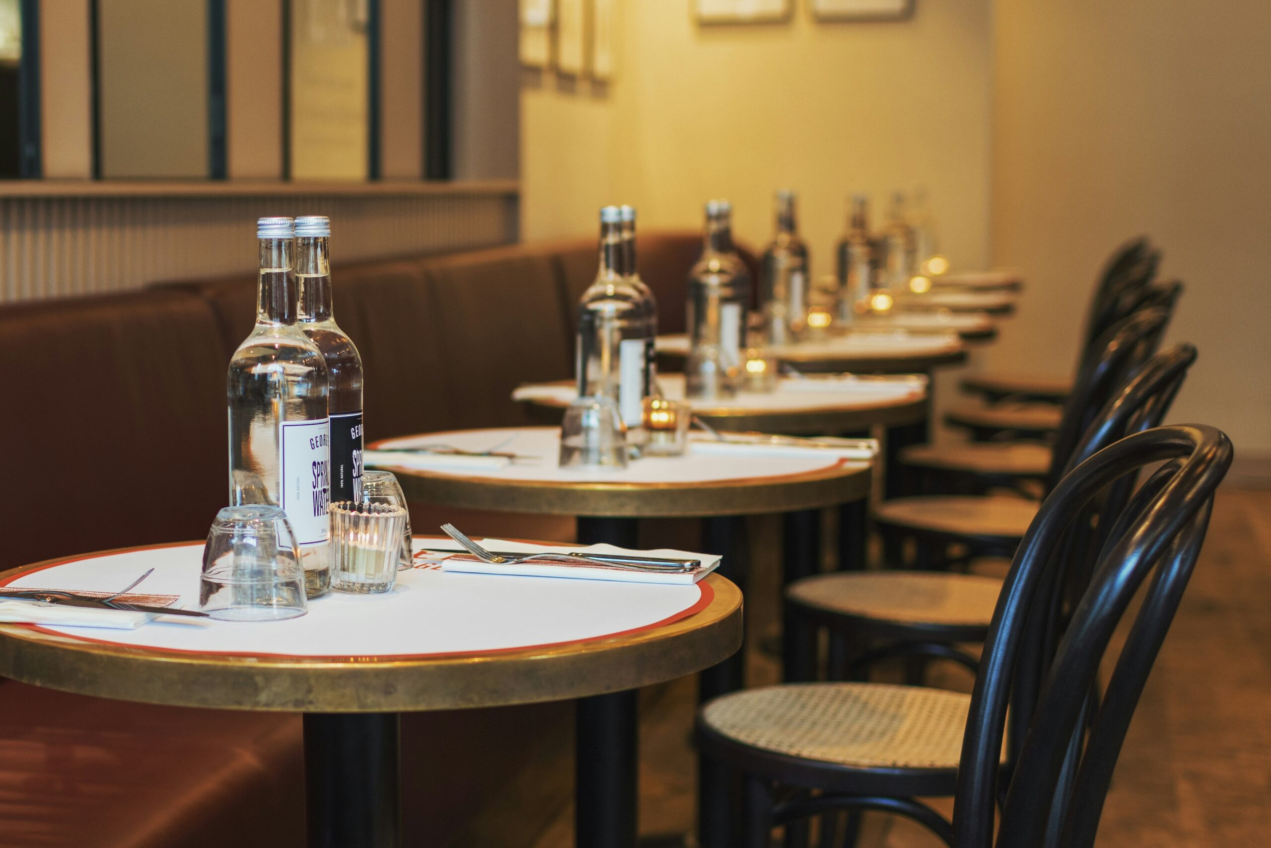 A restaurant with a row of round tables for two, each with a single chair on one side and a long, shared bench on the other. The tables are uniformly set with cups, silverware wrapped in napkins, and two sealed bottles of liquid — a dining experience highlighting why Dallas is worth visiting.