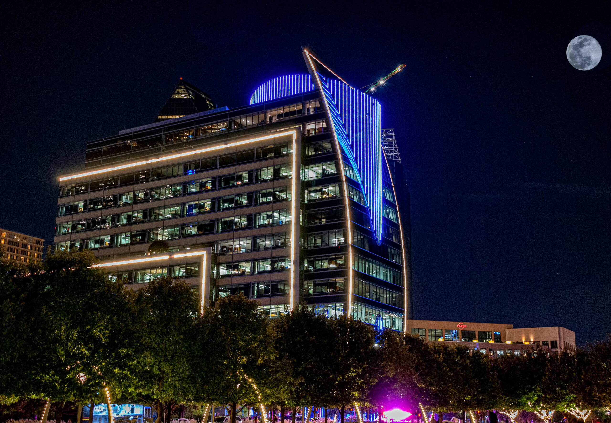 A building highlighted with blue neon lights helps define the Dallas skyline, while a full moon hangs in the sky above and food trucks sit at ground level nestled beneath trees - a beautiful sight and a great option for free things to do in Dallas at night.
