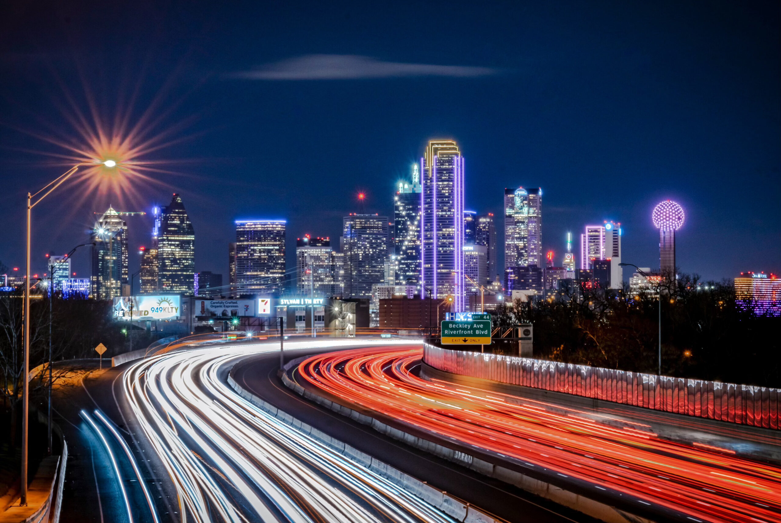 The freeway headed into Dallas from the South is superimposed with lanes of red lights moving into the city and lanes of white lights moving out of Dallas, emphasizing the sense of motion and traffic flow.