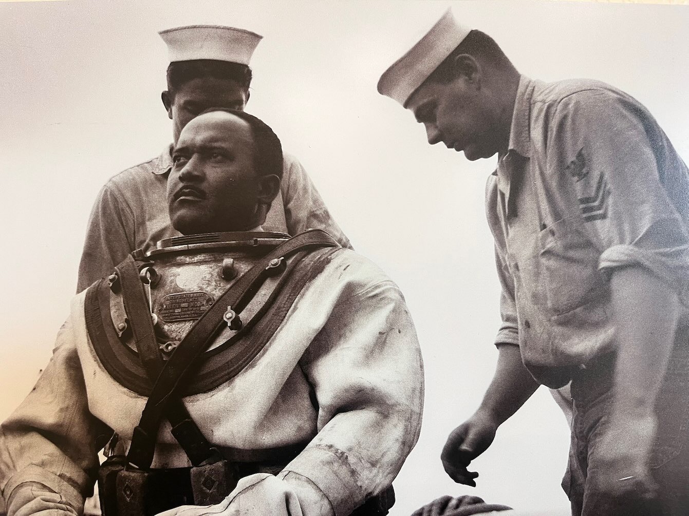 Black U.S. Navy diver in diving gear being assisted by two white sailors preparing for a dive, featured in African American Museum of Dallas reviews.