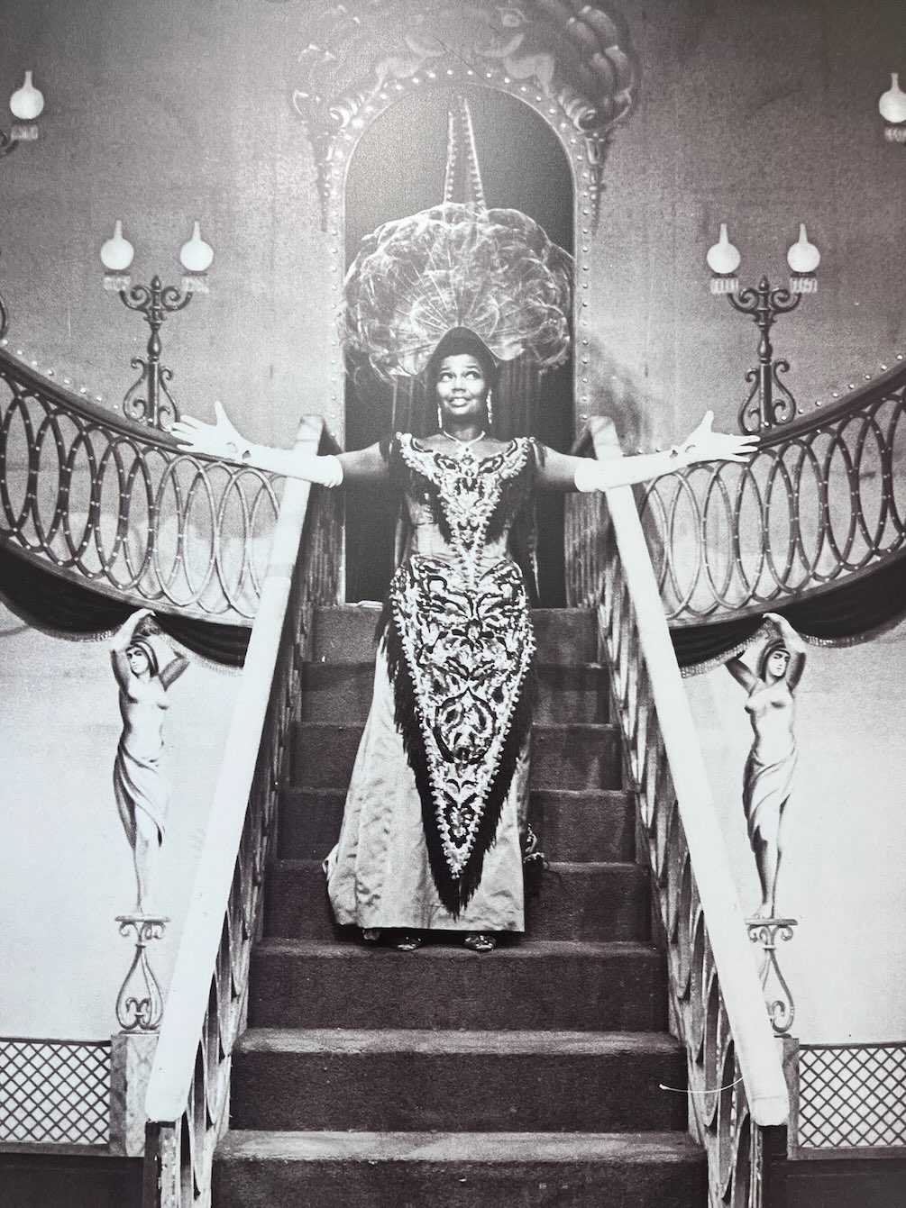 Pearl Bailey descends an ornate staircase with arms spread wide in a theatrical performance pose, featured in African American Museum of Dallas reviews.