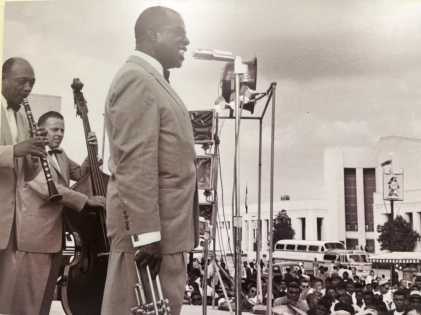 Louis Armstrong singing into a microphone while holding his trumpet as band members play behind him, featured in African American Museum of Dallas reviews.