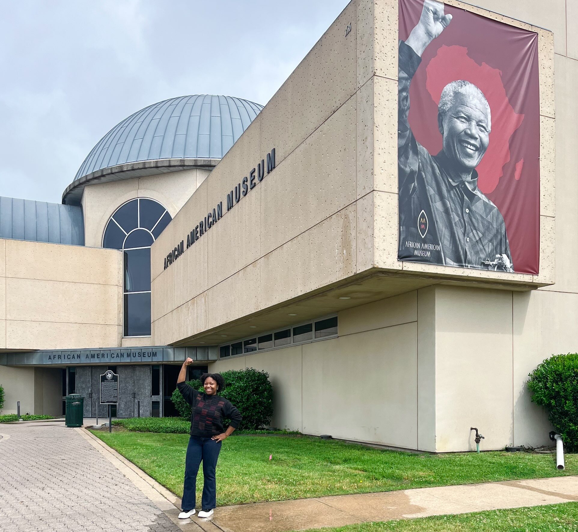 Young woman outside the African American Museum of Dallas raising her arm in a fist, mimicking a Nelson Mandela pose for an upcoming exhibit, as seen in African American Museum of Dallas reviews.