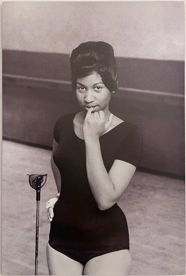 Young Aretha Franklin looks shyly into the camera with finger to lips and hand on hip, featured in African American Museum of Dallas reviews.