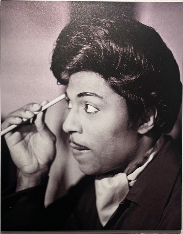Little Richard looks into a mirror while adjusting his hair with a pencil or straight object, featured in African American Museum of Dallas reviews.