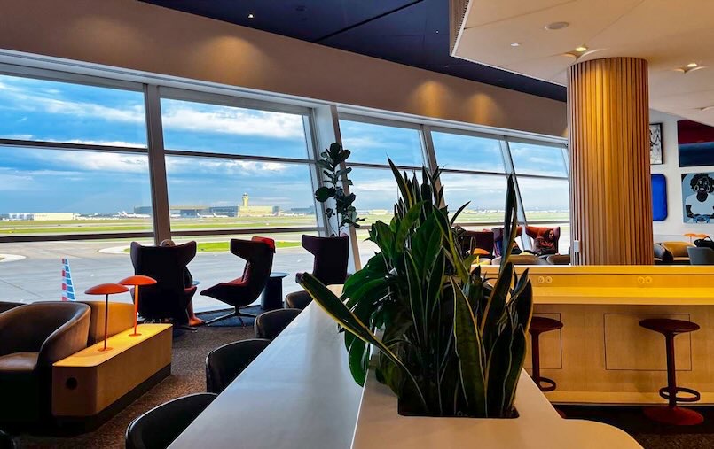 Comfortable seating area inside an airport lounge featuring swivel chairs, armchairs, counter stools, and large windows overlooking the airfield, offering a relaxing space near Dallas airport food and traveler amenities.