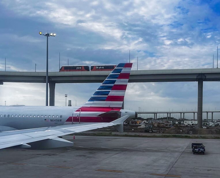 View from the Capital One Lounge at DFW Airport showing the rear of an aircraft on the tarmac, with two Skylink trains traveling on elevated tracks above the airfield, providing convenient access to Dallas airport food options throughout the terminals.