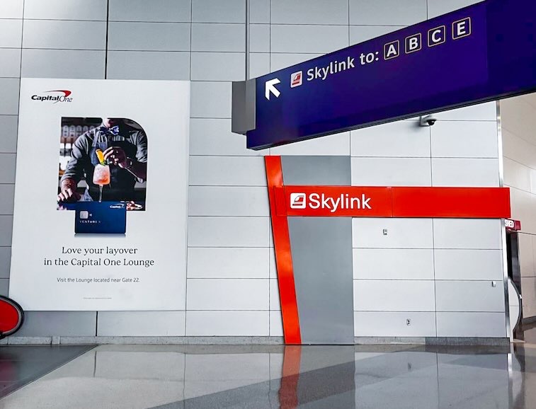 Walkway area near an escalator in a Dallas airport terminal, featuring a Capital One Lounge poster on the wall and Skylink directional signage across from the escalator, near Dallas airport food and traveler amenities.