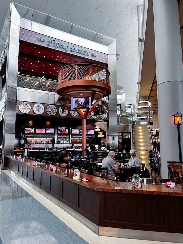 Airport restaurant with an outward-facing dining counter where travelers can enjoy Dallas airport food while watching terminal activity, with a couple seated nearby eating and viewing a television screen.