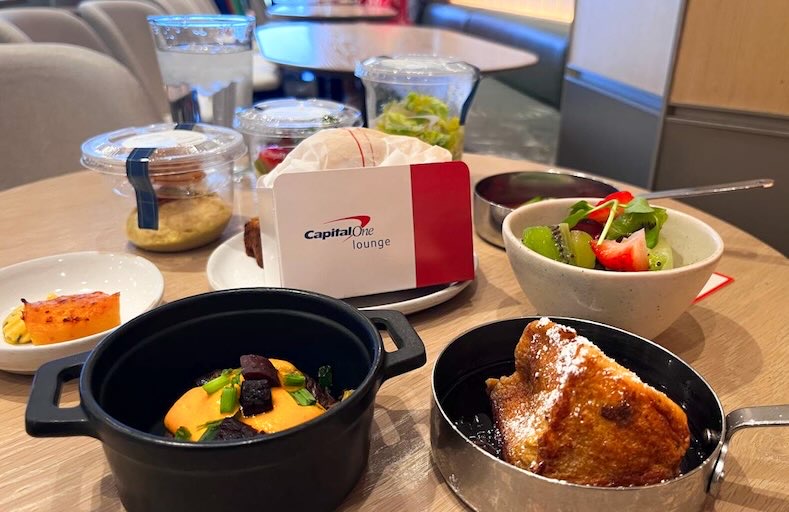 Dining area inside the Capital One Lounge at Dallas airport featuring individually plated complimentary foods, including salads, fresh fruit, vegetables, hot entrées, desserts, house wine, beer, and other beverages available to lounge guests seeking elevated Dallas airport food options.