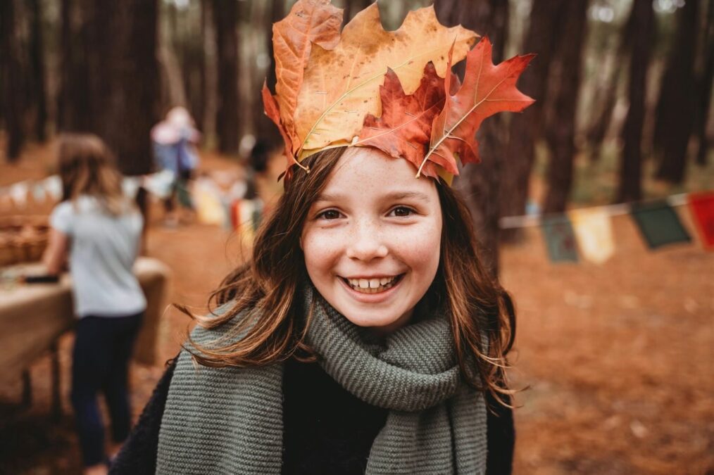 Young girl wearing a fall leaf crown and gray scarf smiles at the camera during a Dallas fall event.
