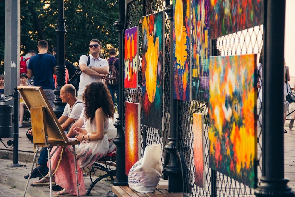 Artist painting at easel with abstract artwork displayed on a fence behind.