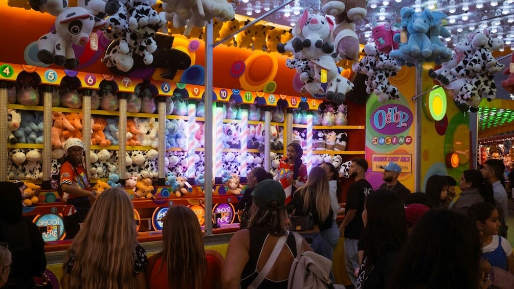 Crowd gathers around a fair game booth with stuffed toy prizes at a Dallas fall event.