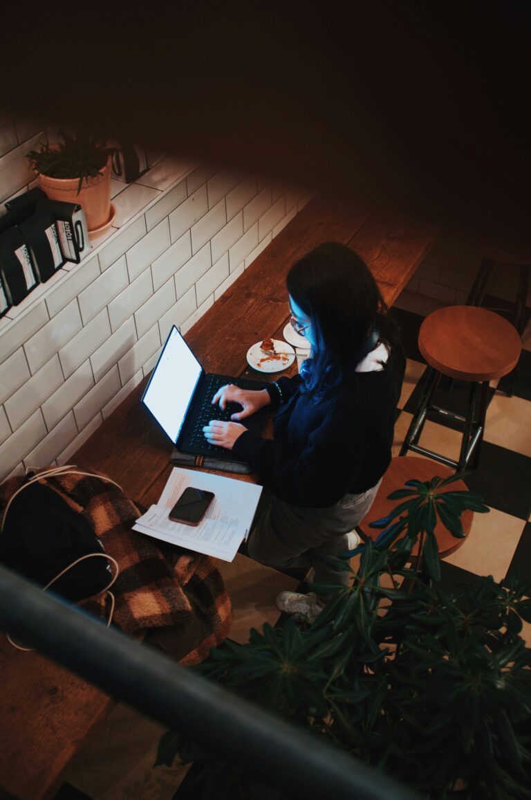 Young woman typing on a laptop at the counter of a late-night coffee shop, her face illuminated by the blue glow of the screen.