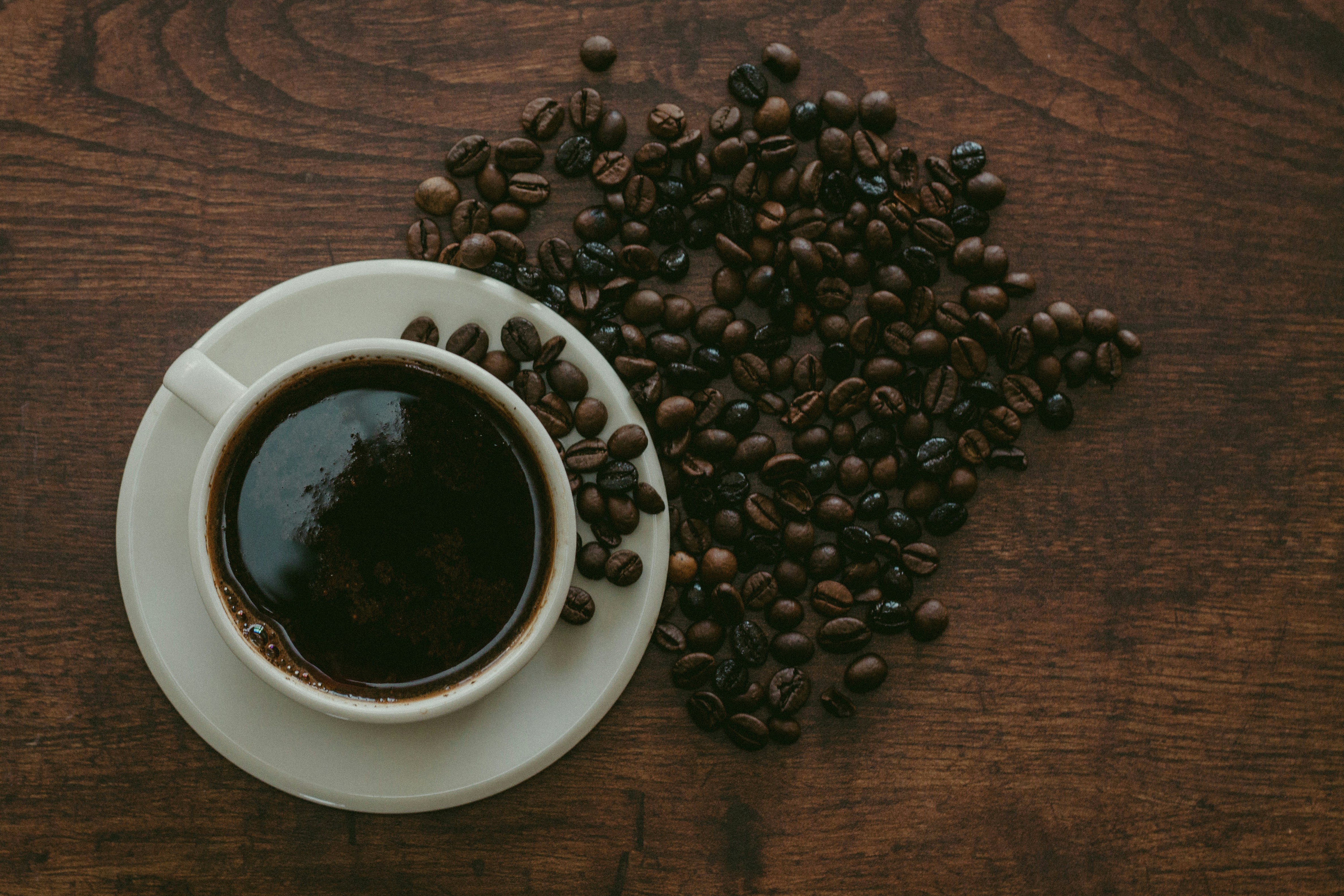 Cup of rich Yemeni coffee on a white saucer with whole coffee beans scattered close on a rippled wooden table.
