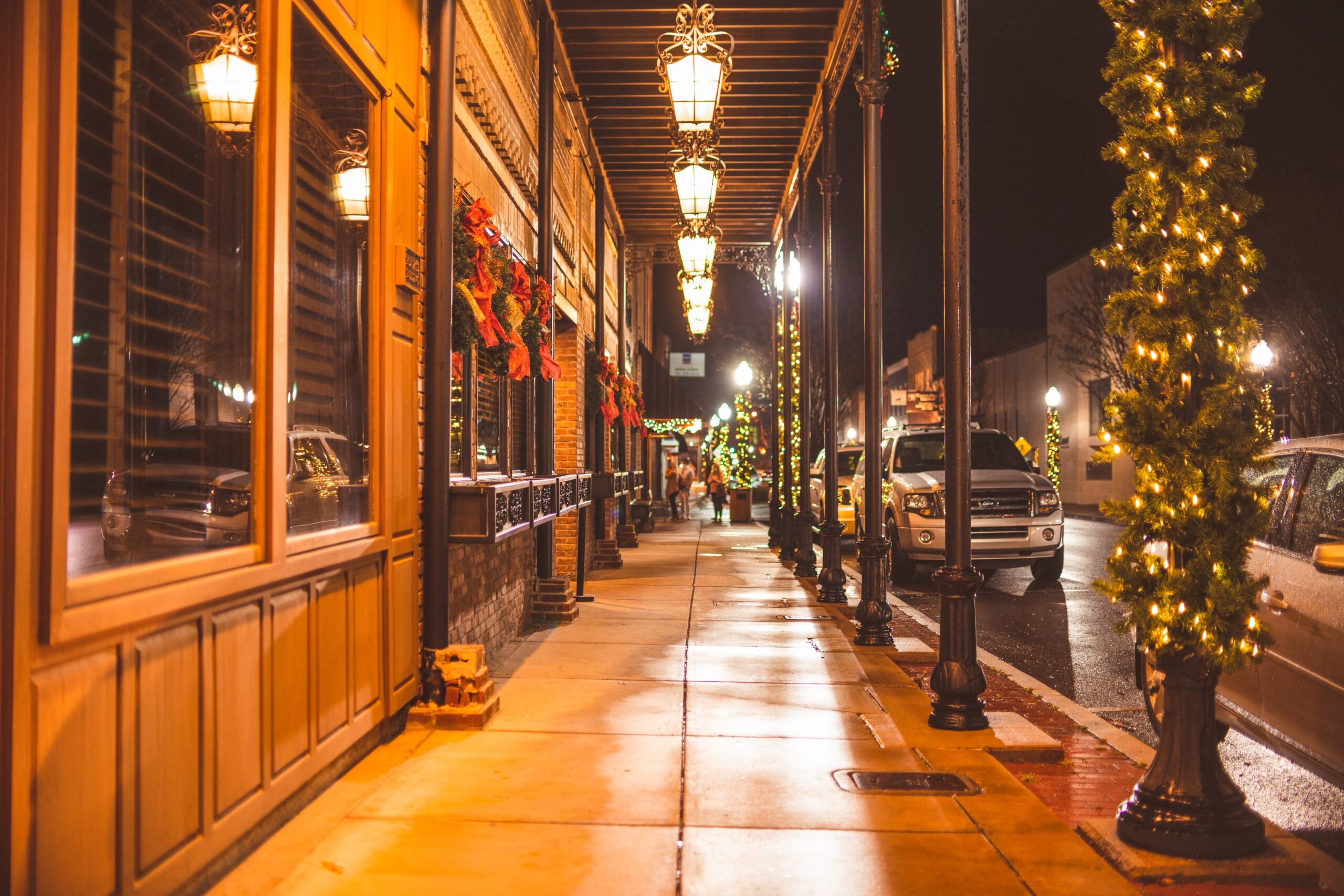 A festively decorated street lined with illuminated shops at night during Christmas in Grapevine, with parked cars and people walking beneath canopy lights.