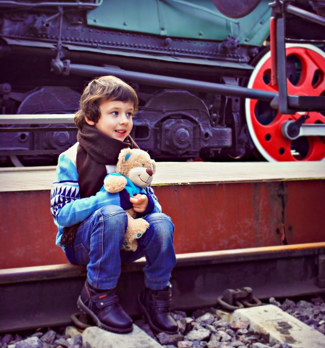 A young boy holding a teddy bear smiles while sitting beside the railroad tracks as he waits for the North Pole Express during Christmas in Grapevine.