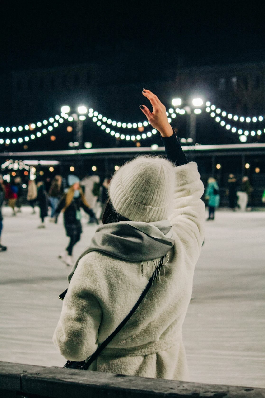 Woman in winter attire waves in recognition toward someone on a crowded ice skating rink during Christmas in Grapevine.