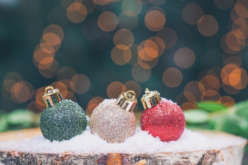 Glittered green, white, and red Christmas ornaments with gold tops arranged in snow on a wooden stump during winter in Dallas.