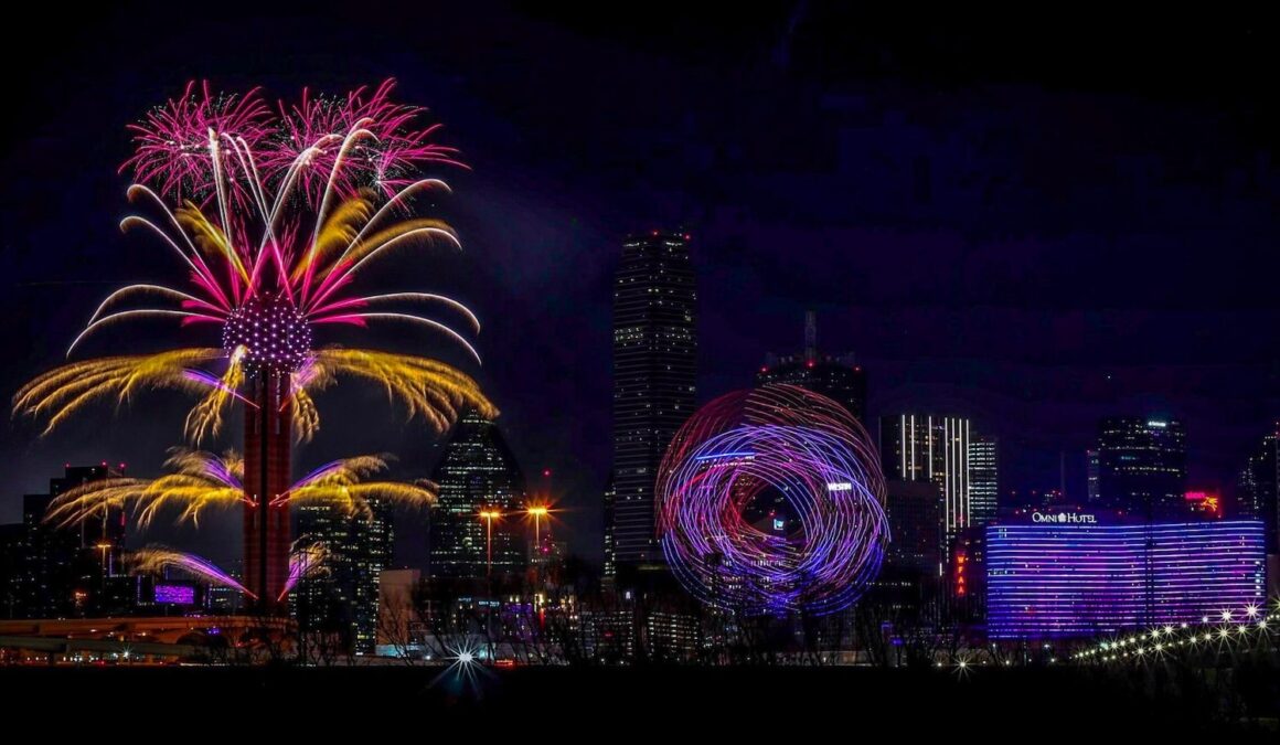 Winter in Dallas night view of Reunion Tower with gold, pink, purple, and white light trails resembling fireworks, with colorful illuminated buildings nearby.