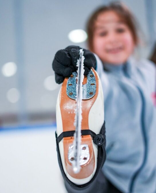 During the winter season, an adult ice skater lifts her foot clearly showing residue along the blade of her boot, and the bottom of her boot with the blade fills the screen. The lady skating holds the blade with a gloved hand while her form is blurred in the background.