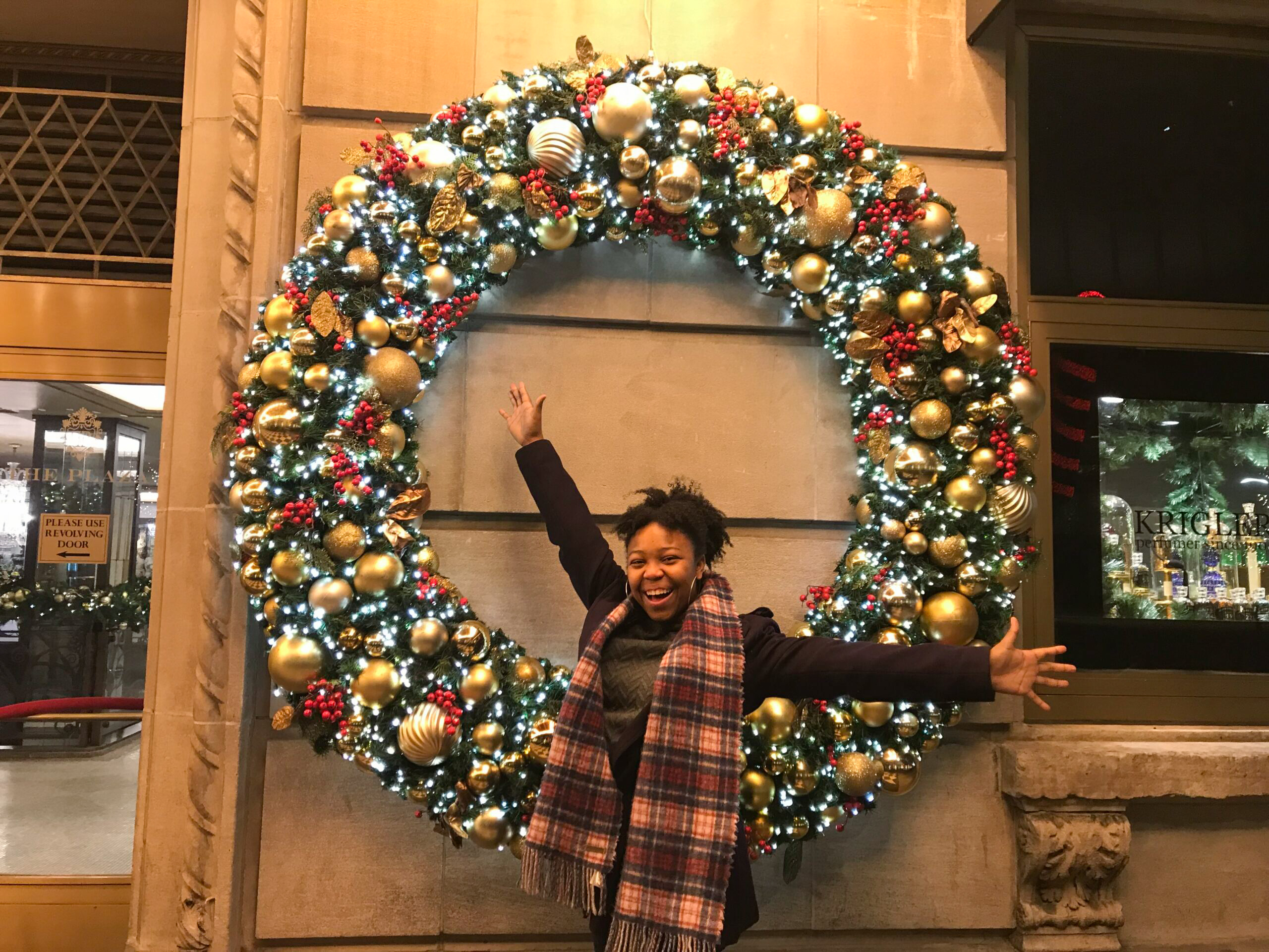 Animated young lady spreading her arms in delight in front of a large green holiday wreath decorated with gold bulbs and red holly during winter in Dallas.