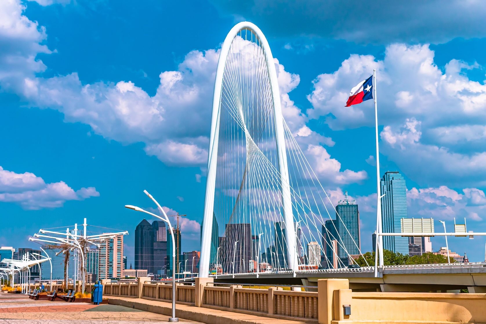 White arched Ronald Kirk Pedestrian Bridge beneath a blue, cloud-dotted sky, with the Dallas skyline in the background and the Texas flag in the foreground.