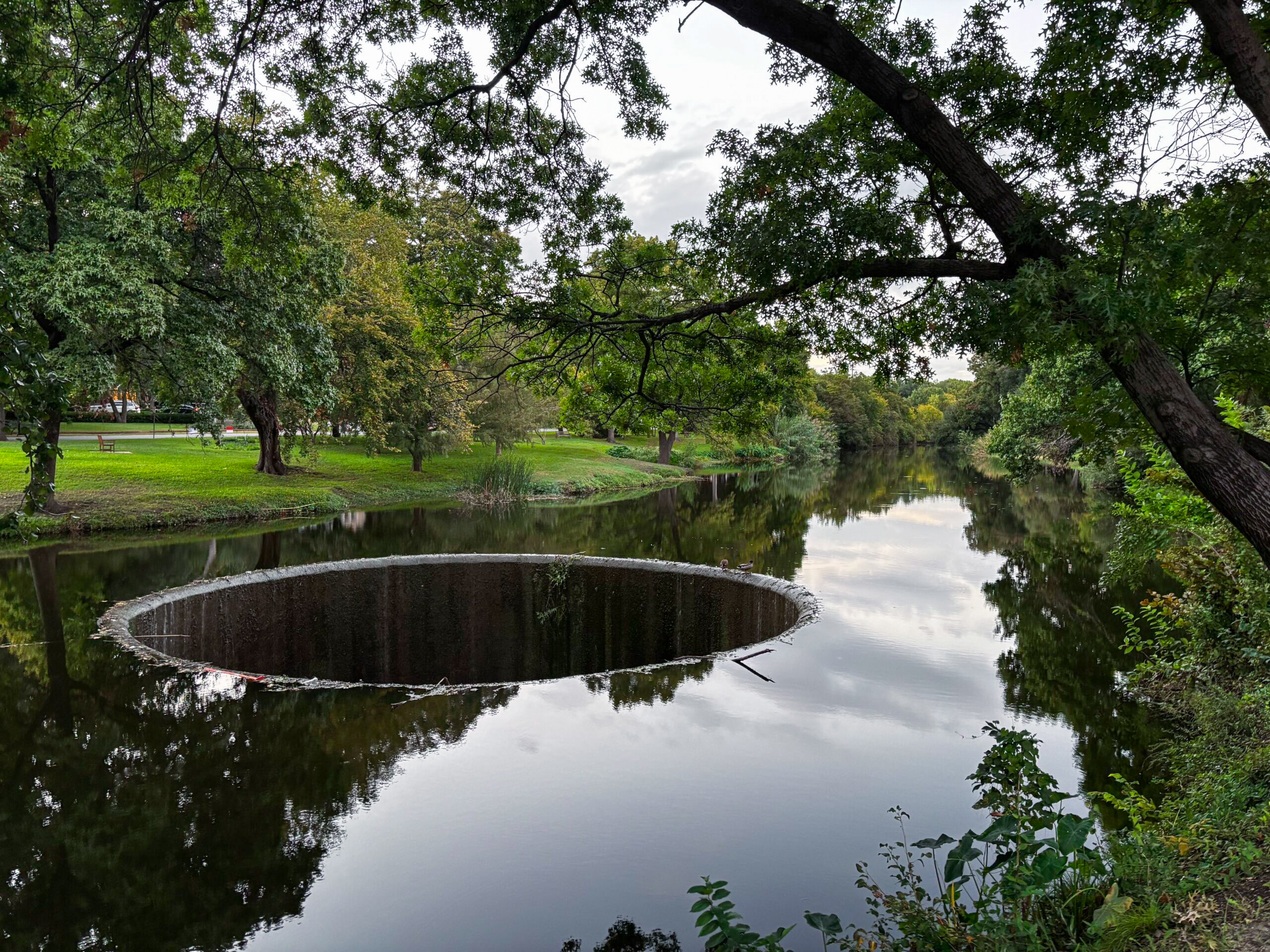 Pond and circular black hole spillway at Connor Park in Highland Park, surrounded by shady trees and green lawns, a peaceful spot for couples seeking free things to do in Dallas.